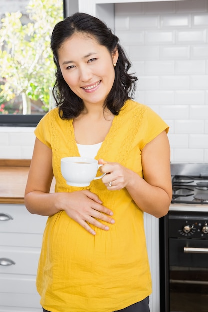 Mujer embarazada sonriente que sostiene la taza y que toca su vientre en la cocina