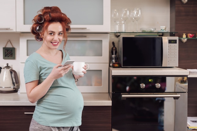 Mujer embarazada sonriente que usa un teléfono inteligente en la cocina