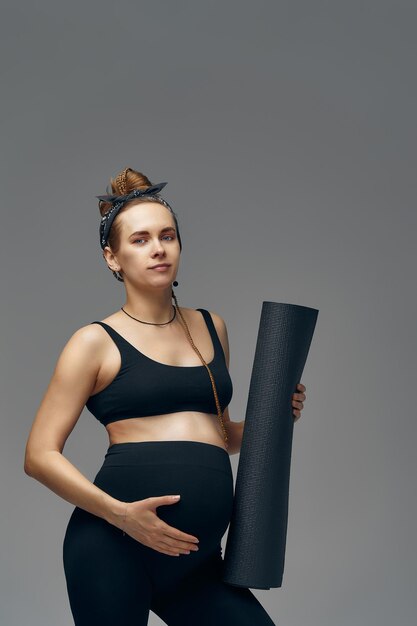 Mujer embarazada sosteniendo una colchoneta de yoga en las manos yendo a hacer deporte mientras espera al bebé mirando sonriendo a la cámara y tocándose el vientre de pie contra la pared gris