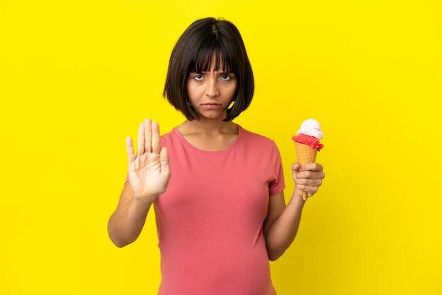 Mujer embarazada sosteniendo un helado de cucurucho aislado sobre fondo amarillo haciendo gesto de parada