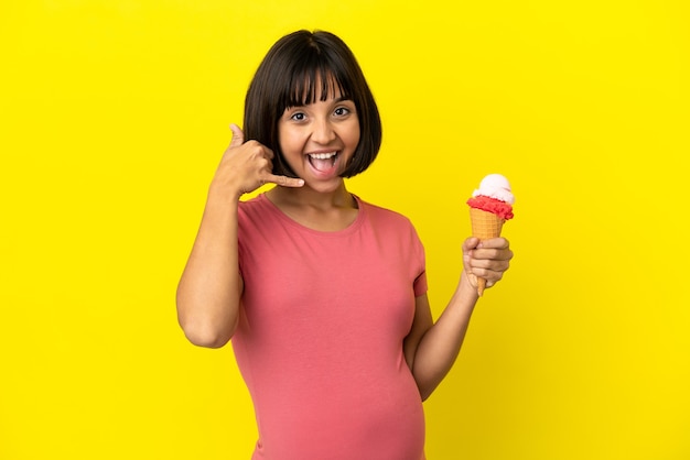 Mujer embarazada sosteniendo un helado de cucurucho aislado sobre fondo amarillo haciendo gesto de teléfono. Llámame señal