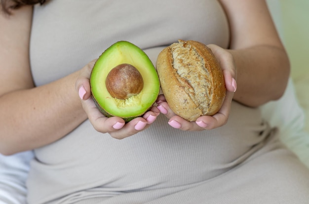 mujer embarazada sosteniendo en la mano la mitad de aguacate con hueso y bollo de pan integral de masa fermentada