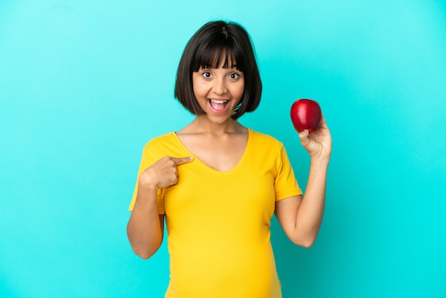 Mujer embarazada sosteniendo una manzana aislada sobre fondo azul con expresión facial sorpresa