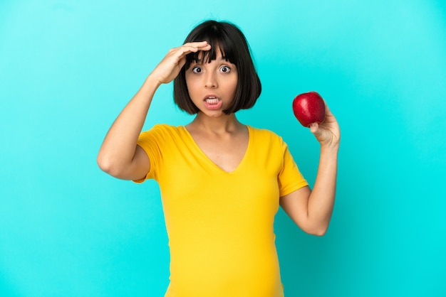 Mujer embarazada sosteniendo una manzana aislada sobre fondo azul haciendo gesto de sorpresa mientras mira hacia el lado