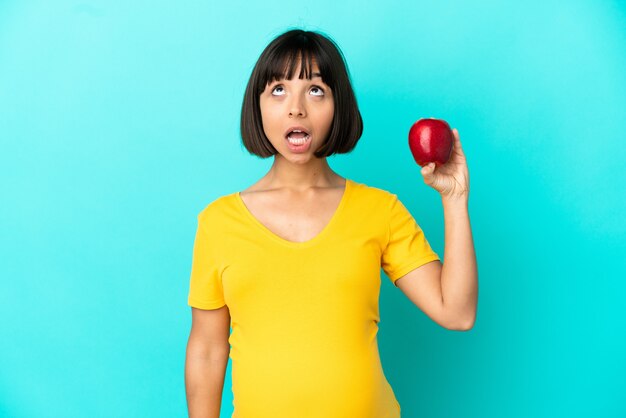 Foto mujer embarazada sosteniendo una manzana aislada sobre fondo azul mirando hacia arriba y con expresión de sorpresa