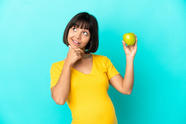 Mujer embarazada sosteniendo una manzana aislada sobre fondo azul teniendo dudas