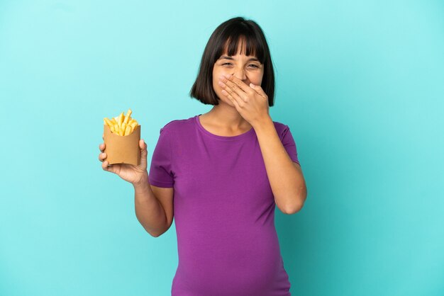 Mujer embarazada sosteniendo patatas fritas sobre fondo aislado feliz y sonriente cubriendo la boca con la mano