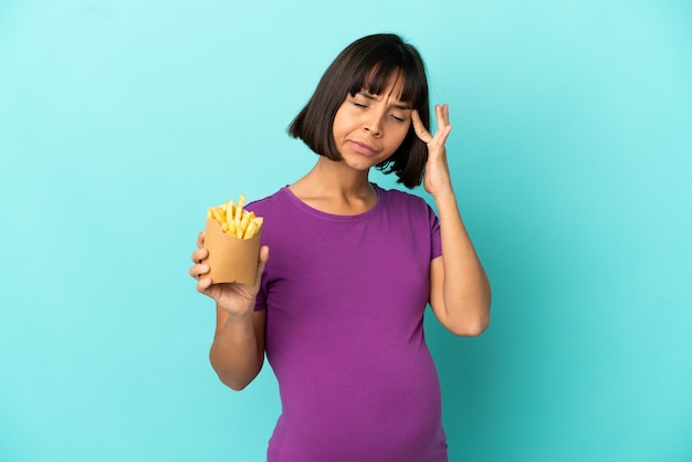 Mujer embarazada sosteniendo patatas fritas sobre pared aislada con dolor de cabeza