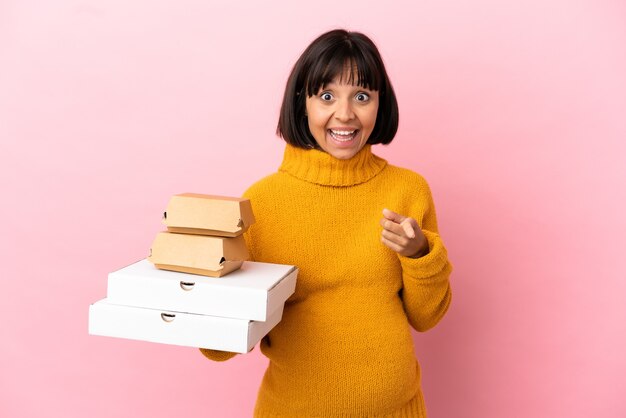 Mujer embarazada sosteniendo pizzas y hamburguesas aisladas sobre fondo rosa sorprendido y apuntando al frente