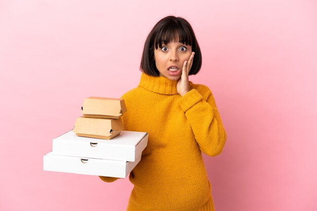 Mujer embarazada sosteniendo pizzas y hamburguesas aisladas sobre fondo rosa con sorpresa y expresión facial conmocionada