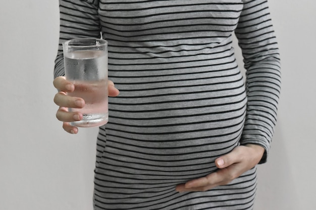 Mujer embarazada sosteniendo un vaso de agua y la mano tocando el vientre sobre fondo gris