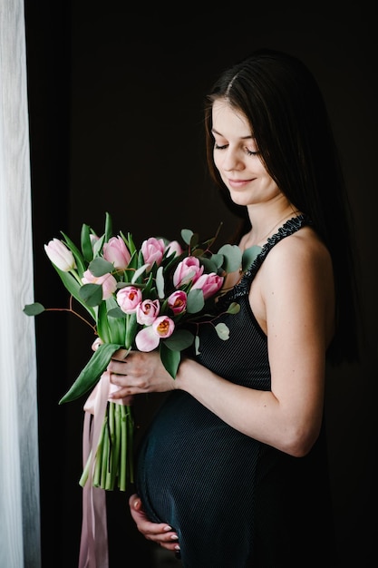 La mujer embarazada sostiene un ramo de flores en las manos, la niña abraza un vientre redondo cerca de la ventana en casa De pie de lado Concepto de maternidad Cerrar nueve meses Baby Shower