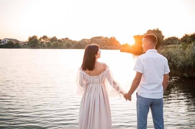 Una mujer embarazada y su esposo se paran en el muelle y se miran un hermoso río y la puesta de sol futuros padres pasan tiempo al aire libre una hermosa joven madre