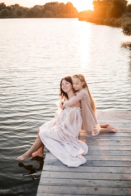 Una mujer embarazada con su hija contra el fondo del agua al atardecer una mujer con barriga disfruta de la puesta de sol junto al río