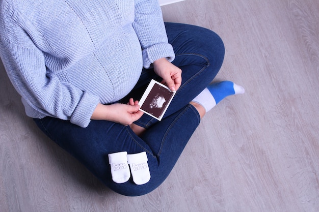 Mujer embarazada en el suéter azul que se sienta en el piso que sostiene la foto del ultrasonido. Mujer embarazada feliz que sostiene imágenes del eco en la mano.