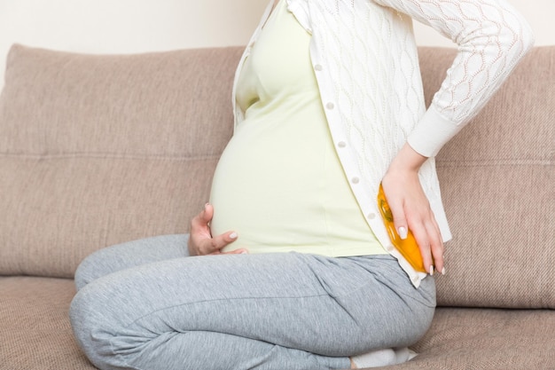 La mujer embarazada sufre de dolor de espalda sostiene una botella caliente o un calentador contra su espalda Concepto de estilo de vida y atención médica de la mujer embarazada