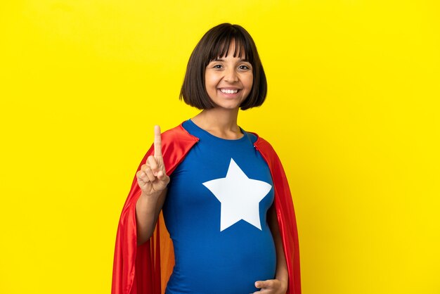 Mujer embarazada superhéroe aislada sobre fondo amarillo mostrando y levantando un dedo