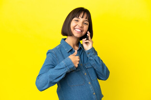 Mujer embarazada con teléfono móvil aislado sobre fondo amarillo dando un pulgar hacia arriba gesto