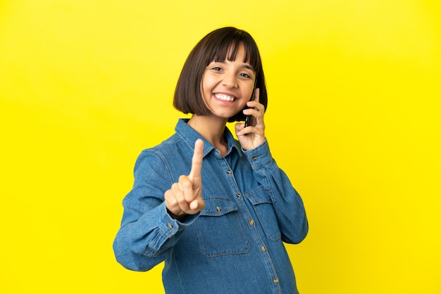 Mujer embarazada con teléfono móvil aislado sobre fondo amarillo mostrando y levantando un dedo