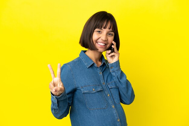 Mujer embarazada con teléfono móvil aislado sobre fondo amarillo sonriendo y mostrando el signo de la victoria