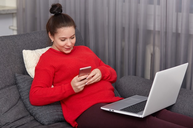 La mujer embarazada tiene el pelo oscuro peinado en moño, escribe mensajes de texto en celulares, navega por las redes sociales