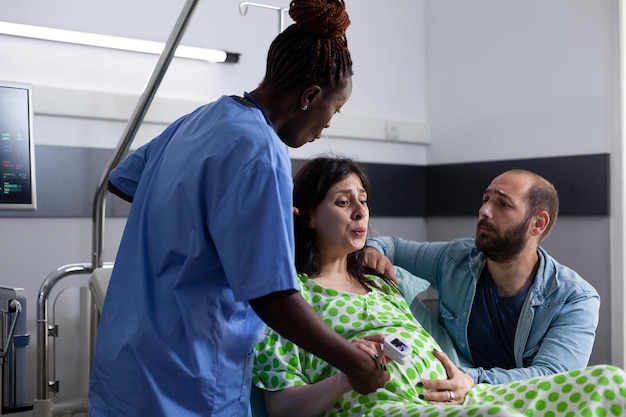 Mujer embarazada con trabajo de parto doloroso acostada en la cama de la sala de hospital, con contracciones. Enfermera afroamericana y esposo apoyando a la futura madre, tomándose de la mano para recibir asistencia médica