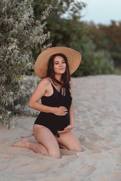 mujer embarazada en traje de baño negro y sombrero en una playa de arena