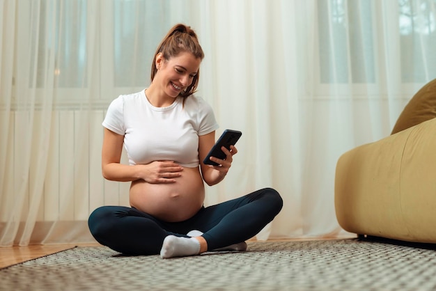 Mujer embarazada usando el teléfono mientras hace ejercicio en una colchoneta de ejercicios en casa