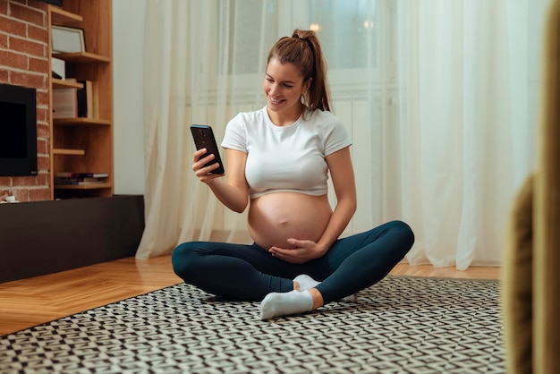 Mujer embarazada usando el teléfono mientras hace ejercicio en una colchoneta de ejercicios en casa