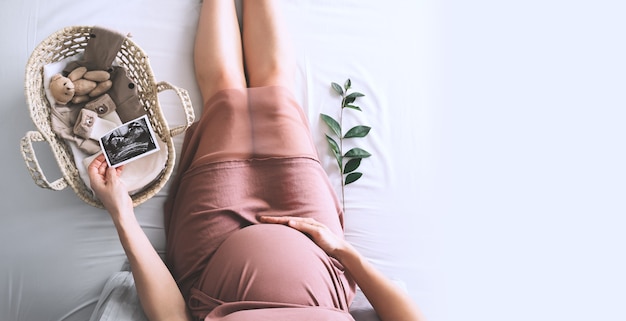 Mujer embarazada vestida con imagen de ultrasonido. Madre con canasta de mimbre de cositas lindas y osito de peluche para recién nacido. Mujer embarazada esperando y preparándose para el parto durante el embarazo.