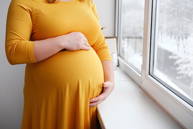 Una mujer embarazada con un vestido amarillo se acaricia el vientre y posa cerca de la ventana Es invierno afuera y está nevando Primer plano del vientre