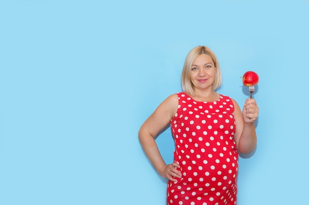 Mujer embarazada con un vestido de lunares rojos sosteniendo una manzana en un tenedor sobre un fondo azul con espacio para copiar