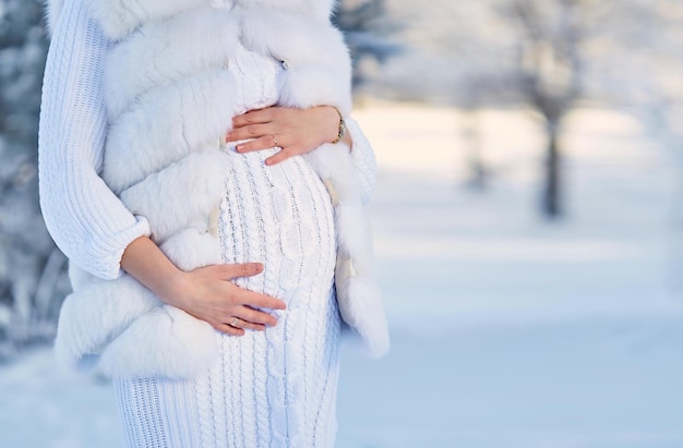 Una mujer embarazada con un vestido de punto blanco y un chaleco de piel en el bosque en invierno