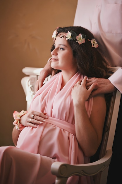 Mujer embarazada en un vestido rosa sentada en una silla detrás de un hombre sonriente de pie