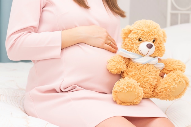 Mujer embarazada en un vestido tiene sus manos sobre su estómago y un oso de juguete.