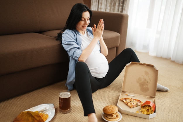 Mujer embarazada con vientre fumando y come alimentos poco saludables en casa. Embarazo, malos hábitos y estilo de vida en el período prenatal. Madre embarazada fea, daño a la salud