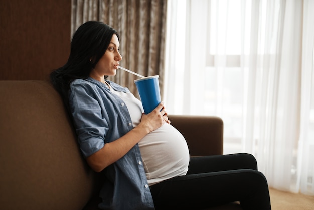 Mujer embarazada con vientre sentado en el sofá y bebe jugo de un vaso grande en casa.