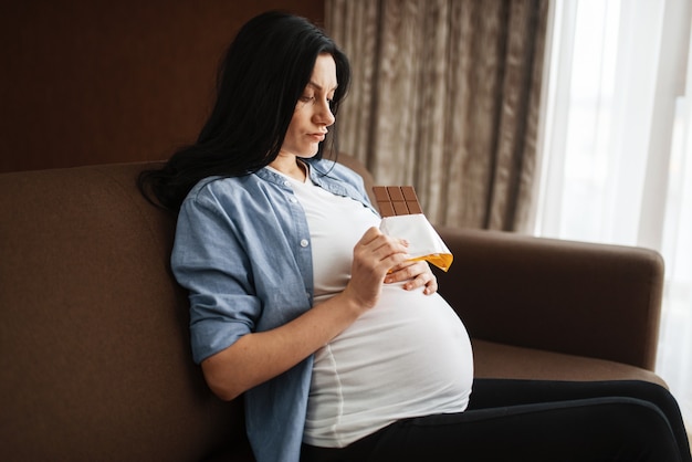 Mujer embarazada con vientre sentado en el sofá y comiendo chocolate. Embarazo, glotonería en período prenatal. Futura mamá descansando en casa