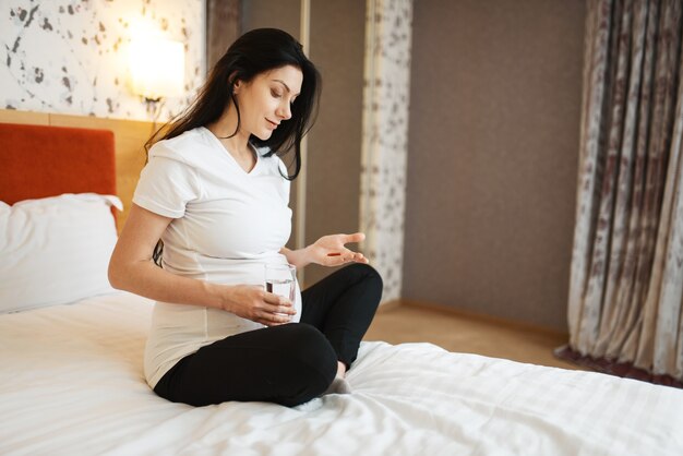 La mujer embarazada con el vientre toma pastillas en la cama en su casa. Embarazo, calma en el período prenatal. Futura mamá descansando en el dormitorio, estilo de vida saludable