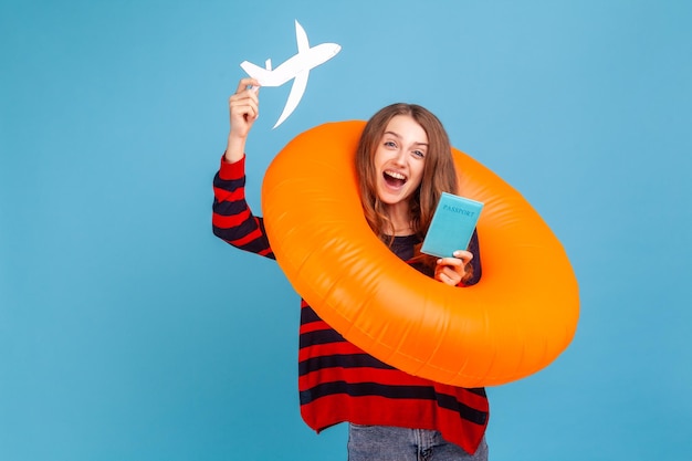 Mujer emocionada con cabello ondulado usando suéter de estilo casual a rayas de pie con pasaporte de anillo de goma y avión de papel feliz de una futura gira Estudio interior aislado en fondo azul