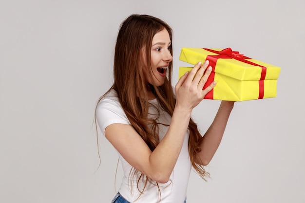 Mujer emocionada con cabello oscuro desempaquetando la caja de regalo mirando dentro con una expresión facial sorprendida regalo de cumpleaños con una camiseta blanca Foto de estudio interior aislada en fondo gris