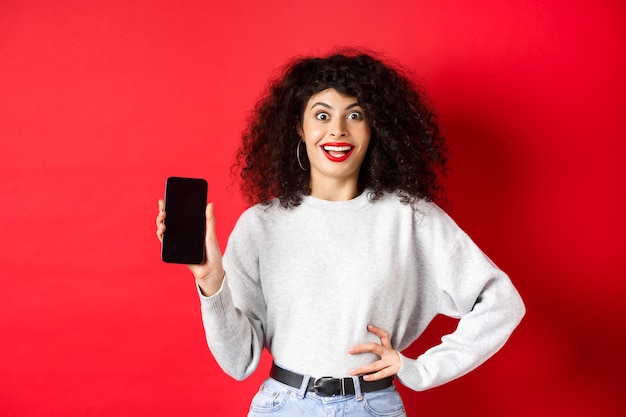 Mujer emocionada con cabello rizado y labios rojos, mostrando la pantalla vacía del teléfono inteligente y gritando de alegría, de pie sobre fondo rojo.