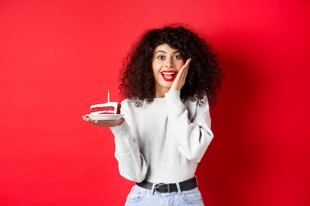 Mujer emocionada celebrando cumpleaños, sosteniendo pastel y mirando sorprendido y feliz, pared roja.