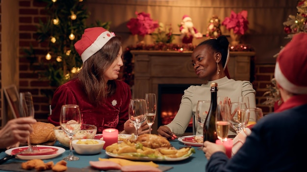 Mujer emocionada discutiendo con un amigo cercano en la cena de Navidad mientras bebe champán. Gente diversa y festiva que celebra la fiesta de invierno con vino espumoso y comida casera tradicional.