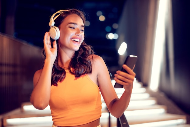 Mujer emocionada disfrutando de su canción favorita durante un paseo por la ciudad por la noche sosteniendo un teléfono celular y sonriendo