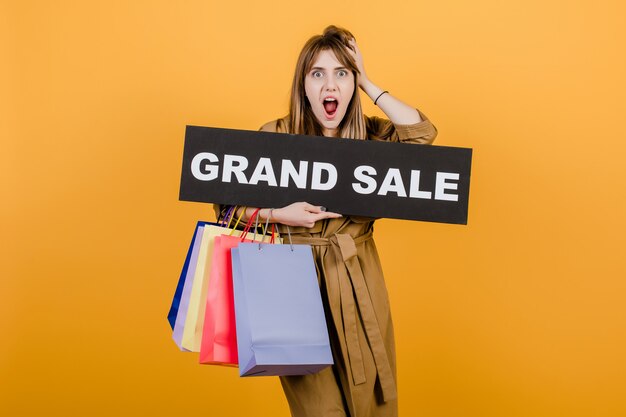 Mujer emocionada hipster en gabardina de otoño con gran cartel de venta y coloridos bolsos de compras