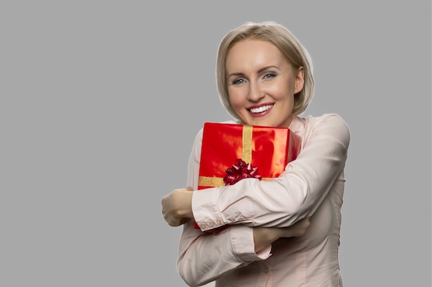 Mujer emocionada joven que sostiene la caja de regalo roja. Mujer alegre con caja de regalo sobre fondo gris. Concepto de regalo de cumpleaños.