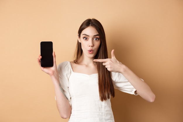 Mujer emocionada mostrando noticias en pantalla, apuntando al teléfono vacío y mirando sorprendido, de pie sobre fondo beige.