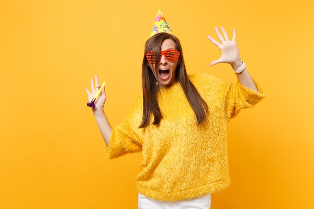 Mujer emocionada en sombrero de cumpleaños de gafas graciosas naranjas con tubo de juego extendiendo las manos, celebrando, disfrutando de vacaciones aisladas sobre fondo amarillo. Personas sinceras emociones, estilo de vida. Área de publicidad.