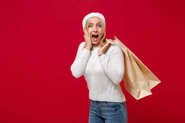 Mujer emocionada con sombrero de suéter blanco aislado de fondo rojo. Concepto de temporada fría de estilo de vida de moda saludable. Simulacros de espacio de copia. Sostenga la bolsa del paquete con las compras después de comprar, ponga la mano en la mejilla.